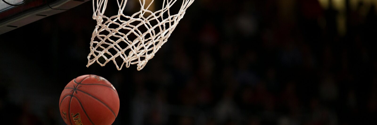 A basketball having just gone through the hoop on an indoor court with spectators in the background.